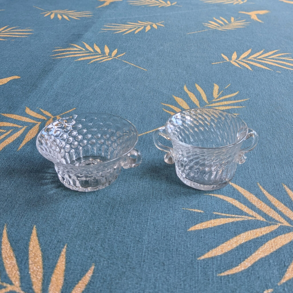 Two small clear textured crystal bowls with handles, standing upright on a blue patterned cloth.