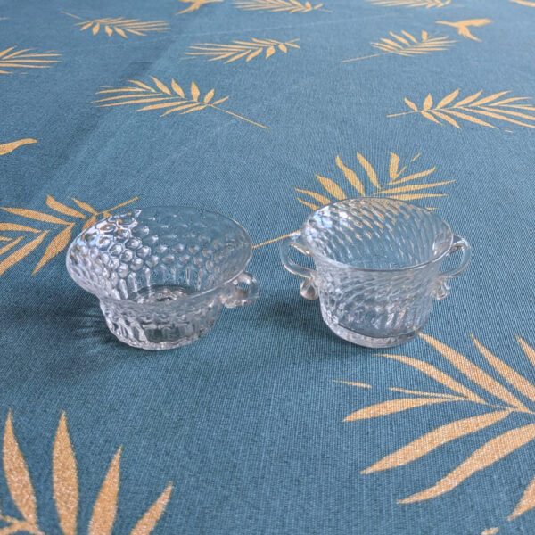 Two small clear textured crystal bowls with handles, standing upright on a blue patterned cloth.
