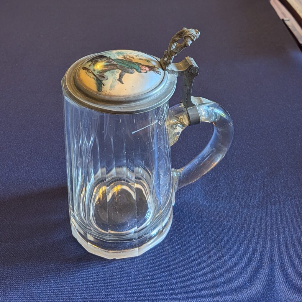 Faceted glass beer tankard with a curved handle and closed illustrated metal lid, three-quarter view.