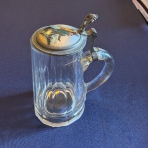Faceted glass beer tankard with a curved handle and closed illustrated metal lid, three-quarter view.