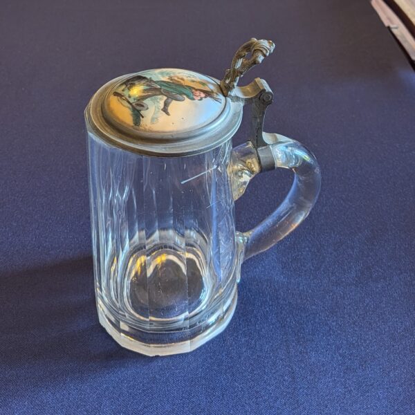Faceted glass beer tankard with a curved handle and closed illustrated metal lid, three-quarter view.