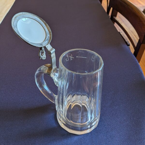Glass beer tankard with the lid raised, showing the rim and the underside of the illustrated insert.