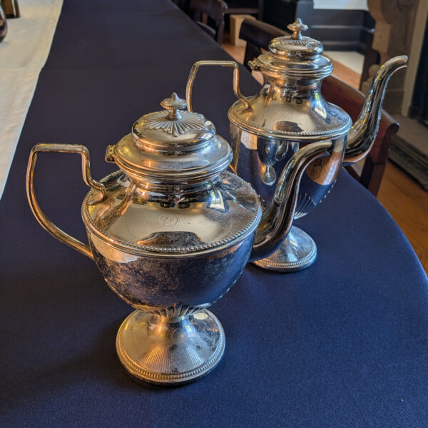 Two silver-coloured metal pots with hinged lids, curved spouts and pedestal bases, showing dark surface discolouration.
