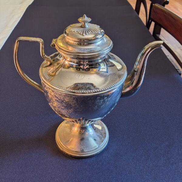 Side view of the shorter silver-coloured metal pot, with its handle, curved spout and closed lid.
