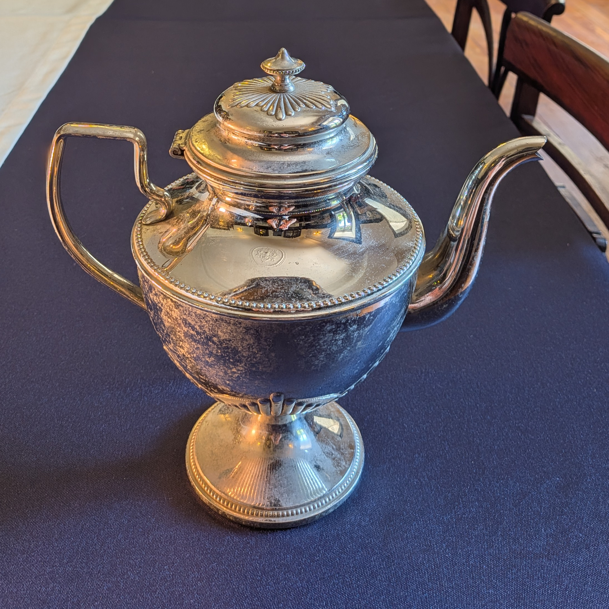 Side view of the shorter silver-coloured metal pot, with its handle, curved spout and closed lid.