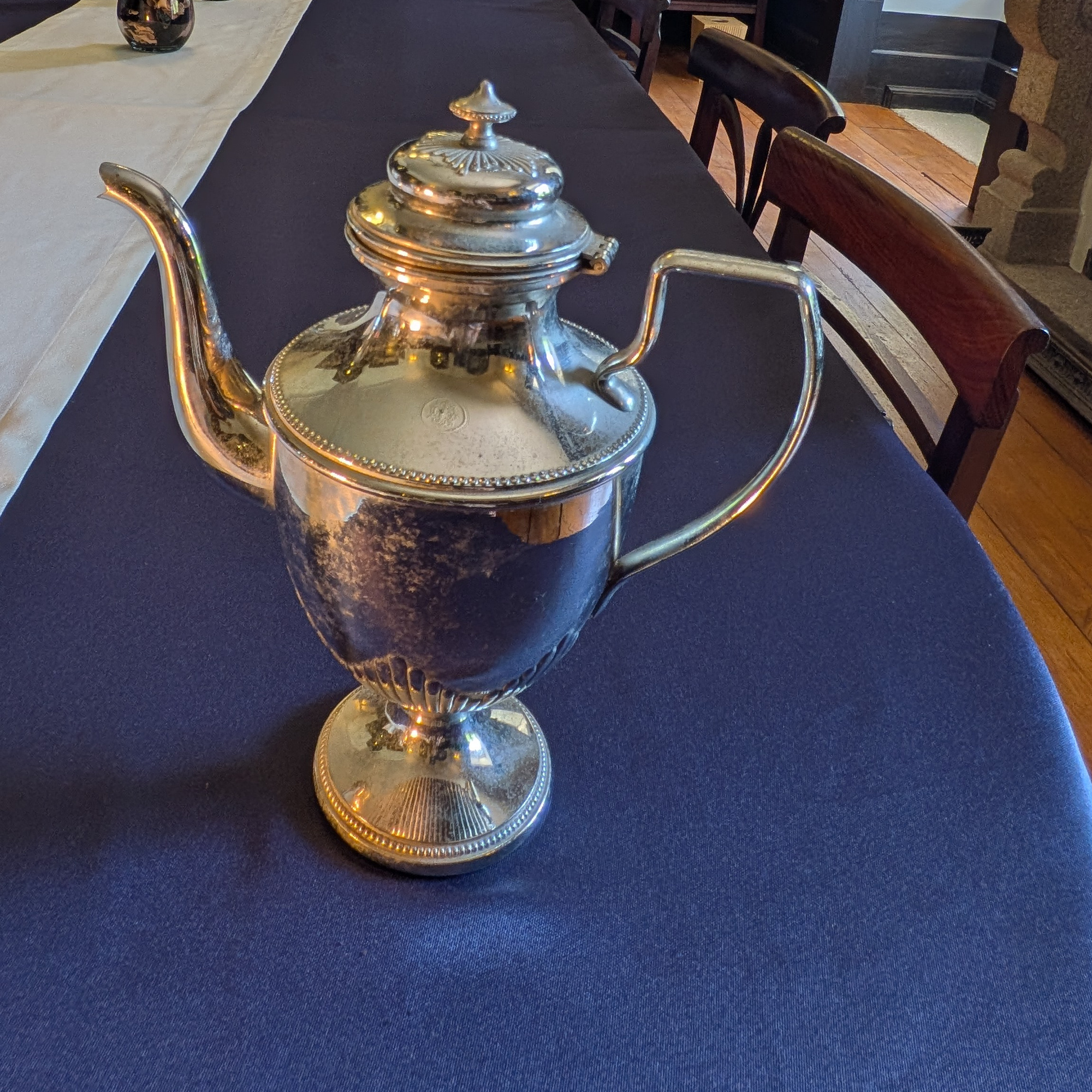 Side view of the taller silver-coloured metal pot, with a closed lid, curved spout and pedestal foot.